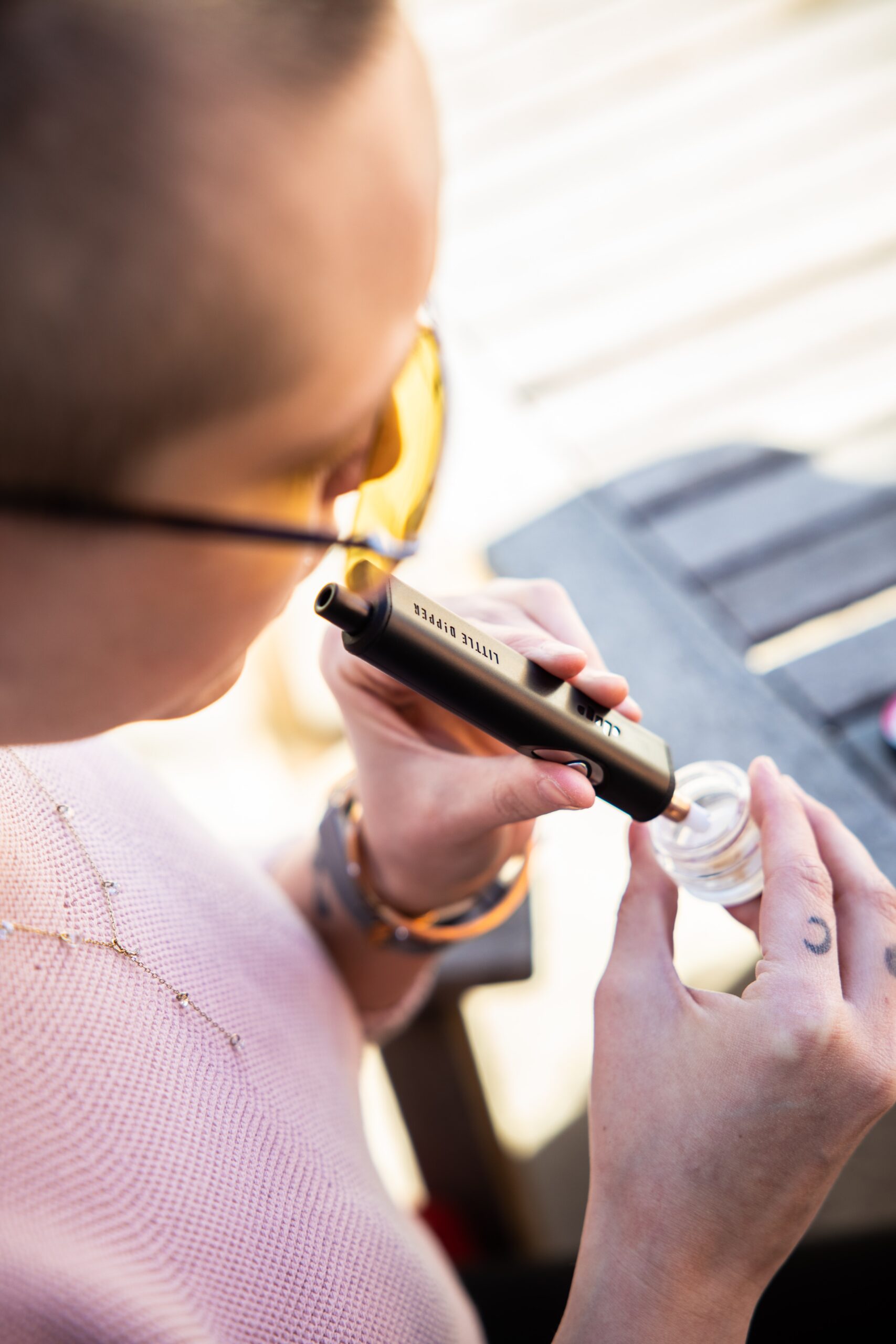 A woman in a pink sweater with hand tattoos and yellow sunglasses enjoys weed rosin using Dip Devices Little Dipper dab straw